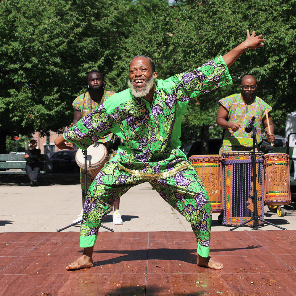 Dancer in a green and purple shirt and pants performing in front of a two percussionists