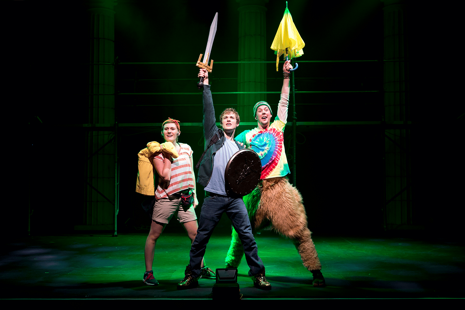 Three performers standing at the front of a stage holding a sword, shield, and umbrella.