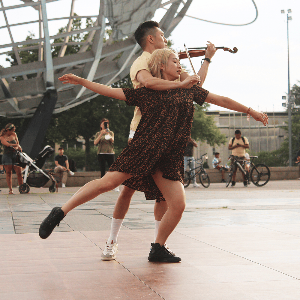 A dancer and a violinist intertwined in performance in front of the Unisphere